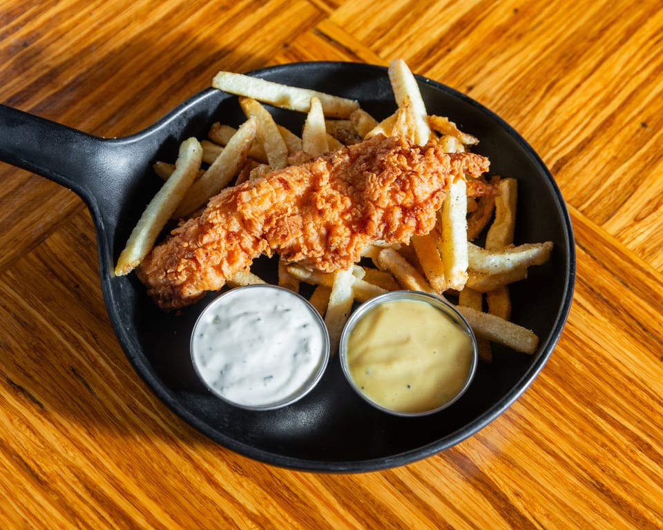 Crispy fried chicken tender and french fries in a black skillet with two dipping sauces.