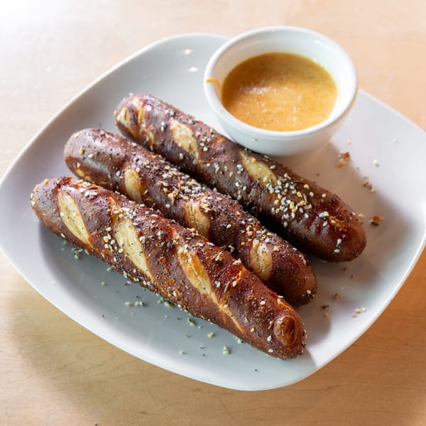 Three pretzel sticks with poppy seeds and sesame seeds, served with a side of mustard dip.