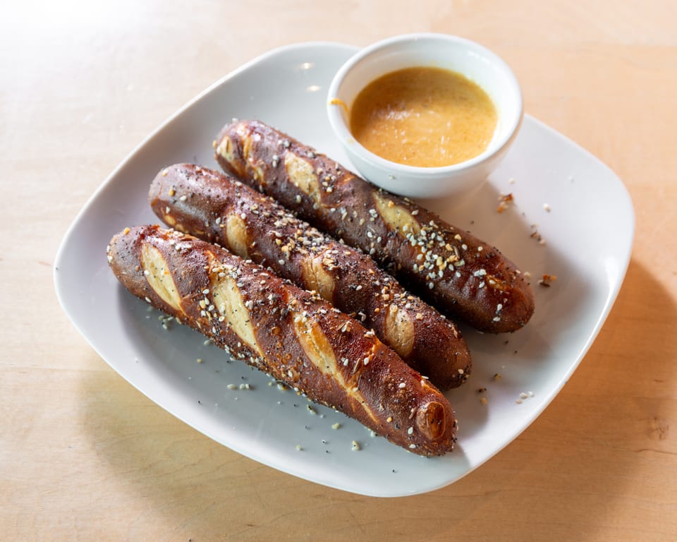 Three pretzel sticks with poppy seeds and sesame seeds, served with a side of mustard dip.