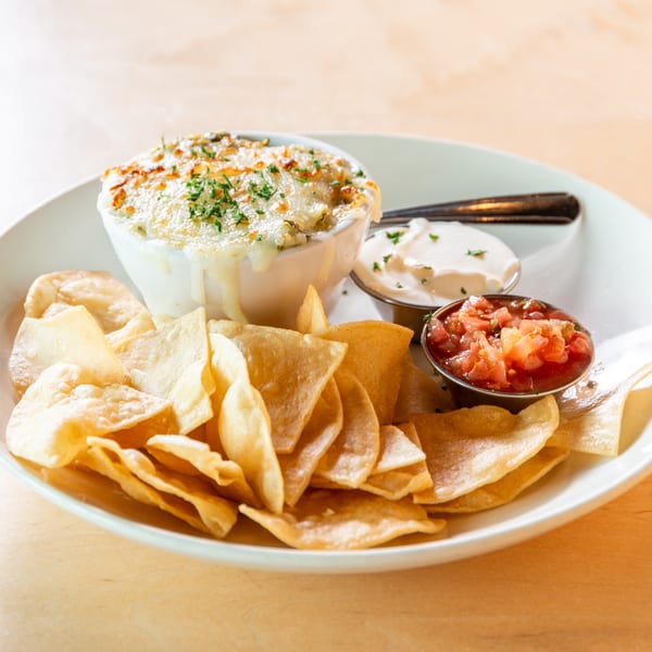 A white bowl of cheesy dip with tortilla chips, salsa, and sour cream on a wooden table.