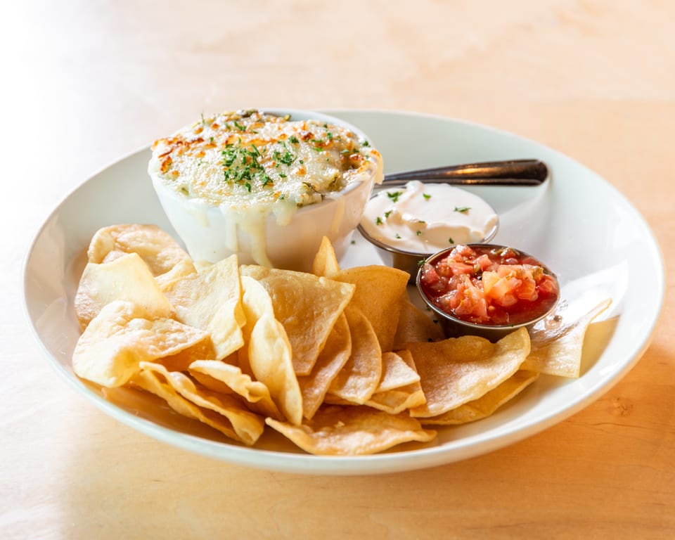 A white bowl of cheesy dip with tortilla chips, salsa, and sour cream on a wooden table.