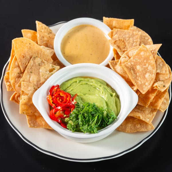 Crispy tortilla chips surround bowls of guacamole with red peppers and cheese dip on a white plate.