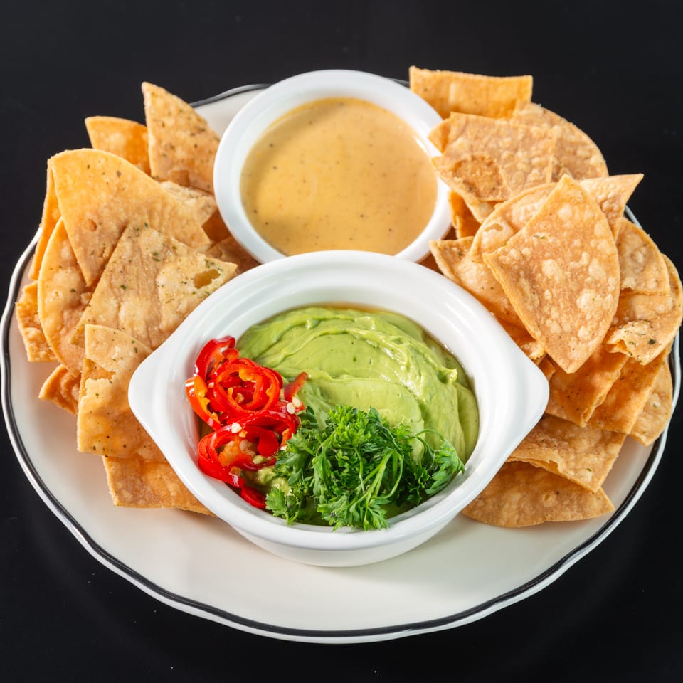 Crispy tortilla chips surround bowls of guacamole with red peppers and cheese dip on a white plate.