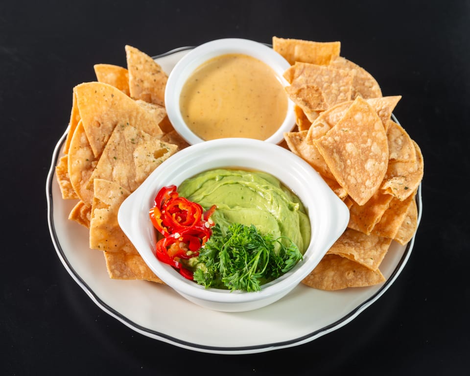 Crispy tortilla chips surround bowls of guacamole with red peppers and cheese dip on a white plate.