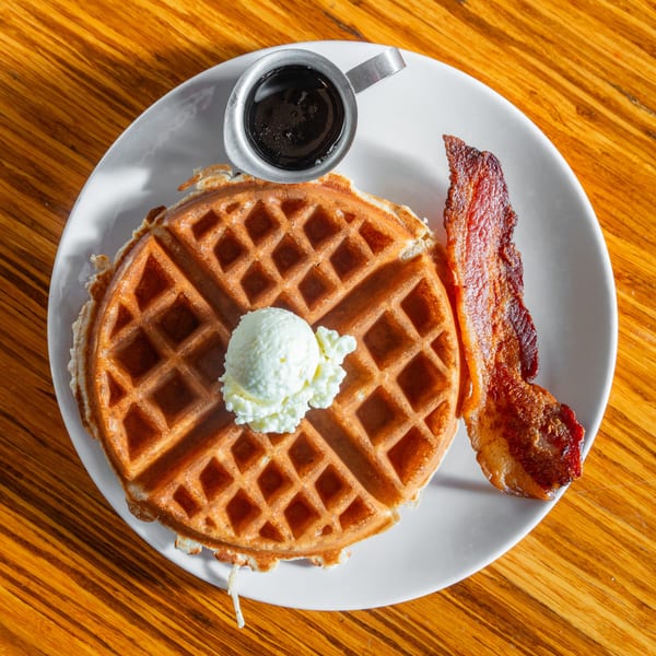 Golden waffle with butter, crispy bacon, and a side of syrup on a white plate.