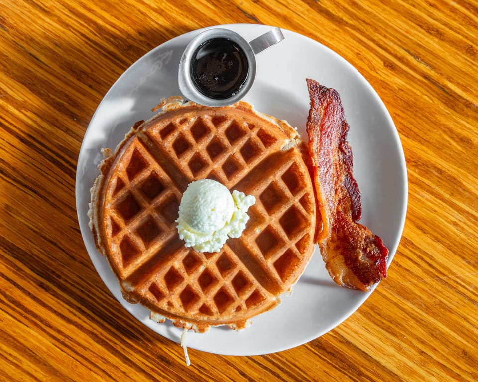 Golden waffle with butter, crispy bacon, and a side of syrup on a white plate.
