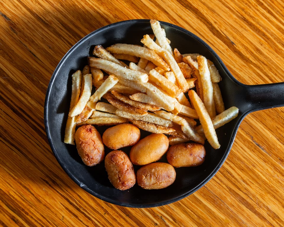 Golden french fries and several mini corn dogs in a black serving dish on a wooden table.