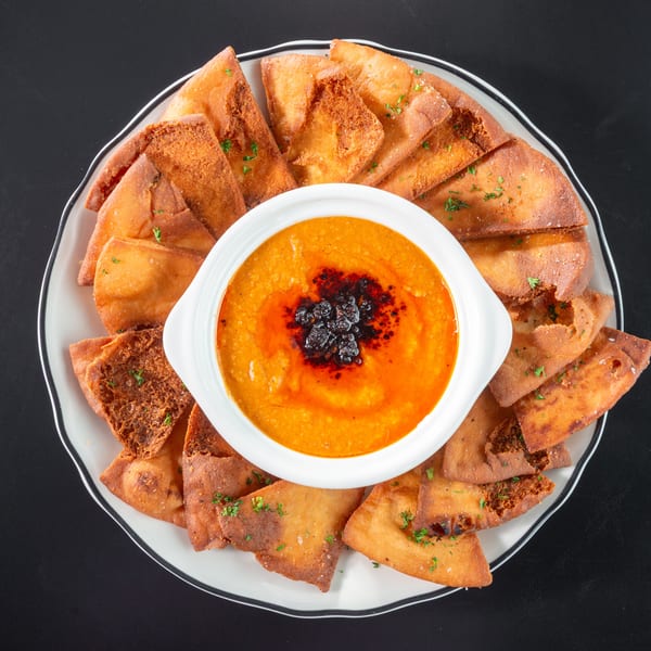 Overhead view of a white bowl of orange dip with dark red oil, surrounded by pita bread.