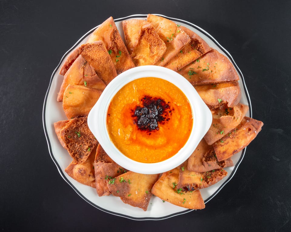 Overhead view of a white bowl of orange dip with dark red oil, surrounded by pita bread.