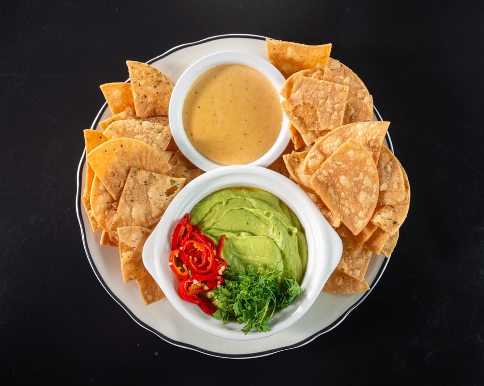 A plate of tortilla chips with bowls of guacamole, queso, and sliced red peppers.